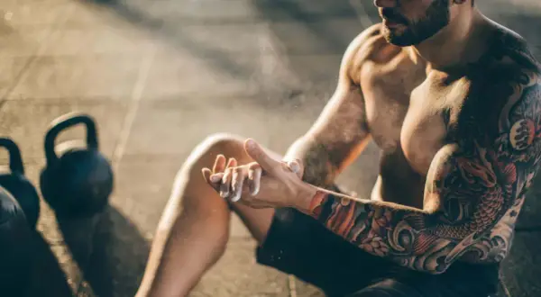 Ein Mann mit beeindruckenden herren tattoos bereitet sich mit Magnesium vor. Kettlebells liegen im Hintergrund.