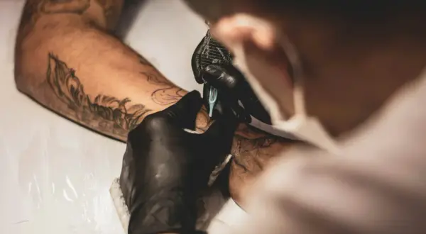 Tätowierer mit schwarzen Handschuhen arbeitet an einem Arm. Ein detailliertes Tattoo ohne Schattierung entsteht.