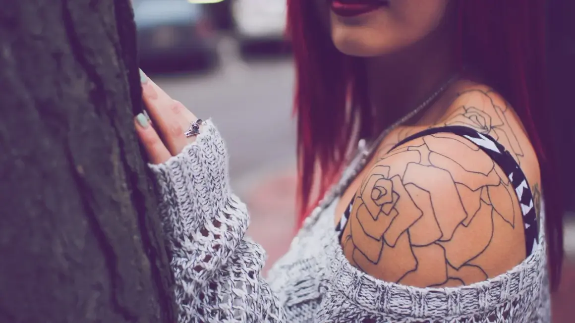 Dezent blühendes Tattoo Schulter Frau, eine zarte Rose ziert ihre Haut, umrahmt von einem Strickpullover.