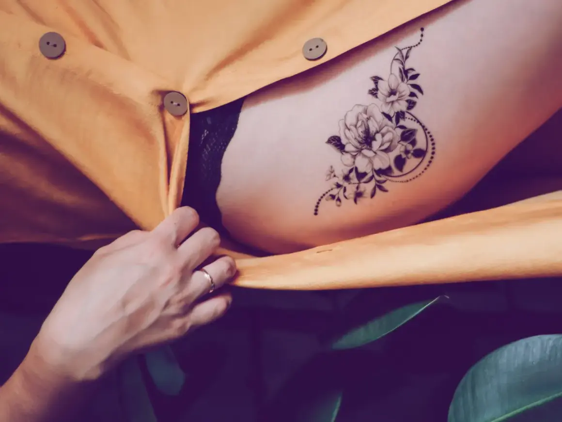Ein zartes Tatoo mit einer Blume ziert die Haut, umrahmt von einem gelben Stoff und grünen Blättern.