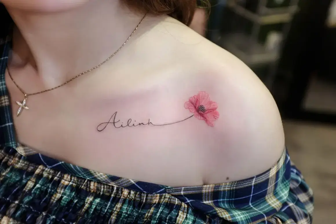 Ein zartes Tattoo mit dem Namen "Ailinh" und einer roten Mohnblume auf der Schulter.