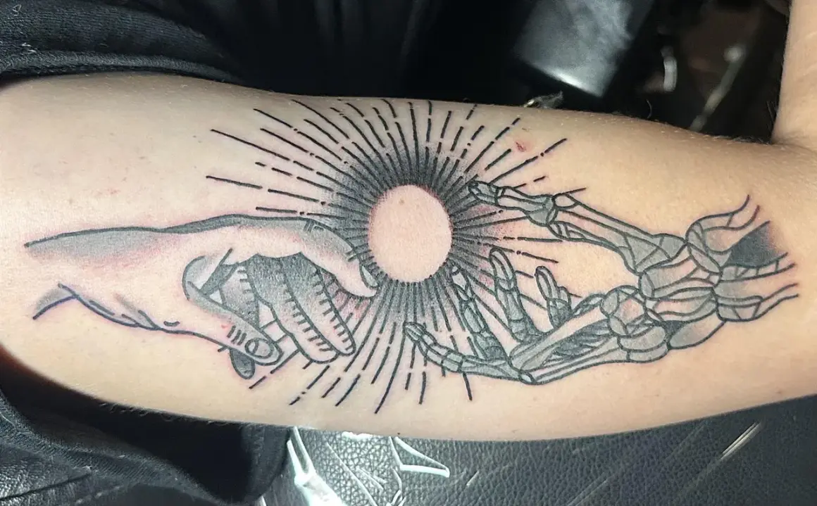Ein Tattoo zeigt eine menschliche Hand und eine Skeletthand, die sich auf einen hellen Kreis zubewegen. Die Haut ist leicht gerötet, als ob sie gerade erst aufgerissen wurde, um das Kunstwerk zu enthüllen.