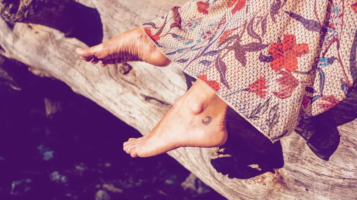 Ein Fuß mit einem kleinen Mond-Tattoo ruht auf einem Baumstamm, bedeckt von einem gemusterten Stoff.