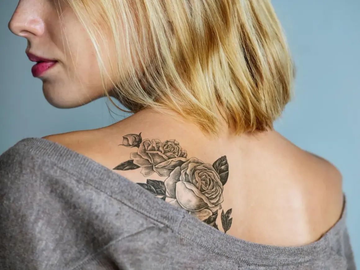 Ein frisches Tattoo mit Rosen auf der Schulter einer Frau, das mit einem grauen Pullover abgedeckt wird.