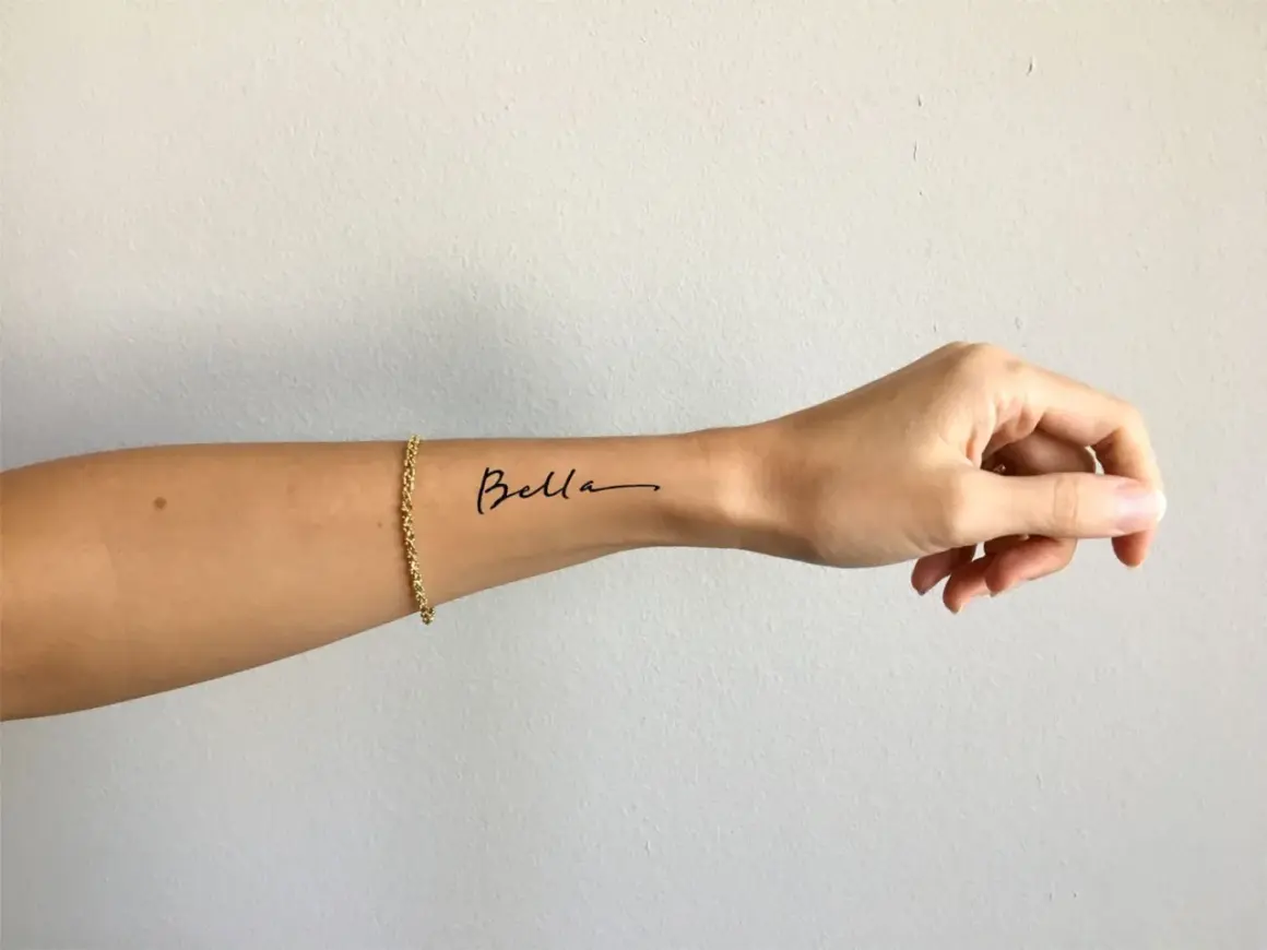 Ein zartes Tattoo-Armband mit dem Namen "Bella" auf dem Handgelenk, geschmückt mit einer goldenen Kette.