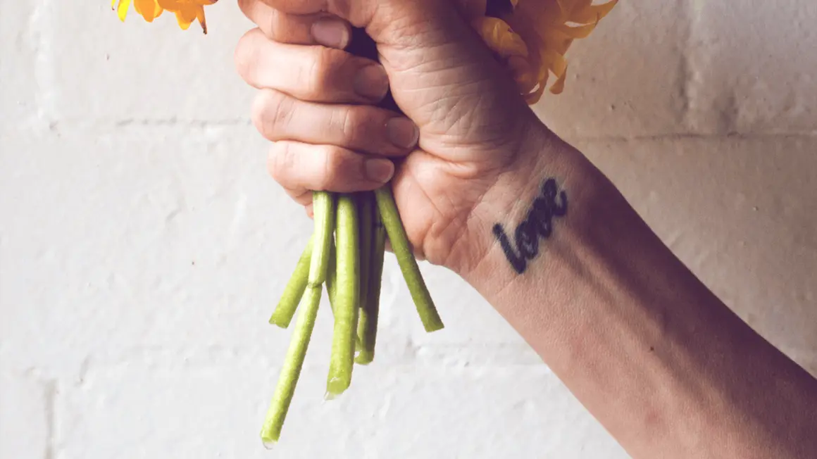 Ein Handgelenk mit einem "Love"-Tattoo, das an ein Freundschaftstattoo erinnert, hält einen Strauß gelber Blumen.