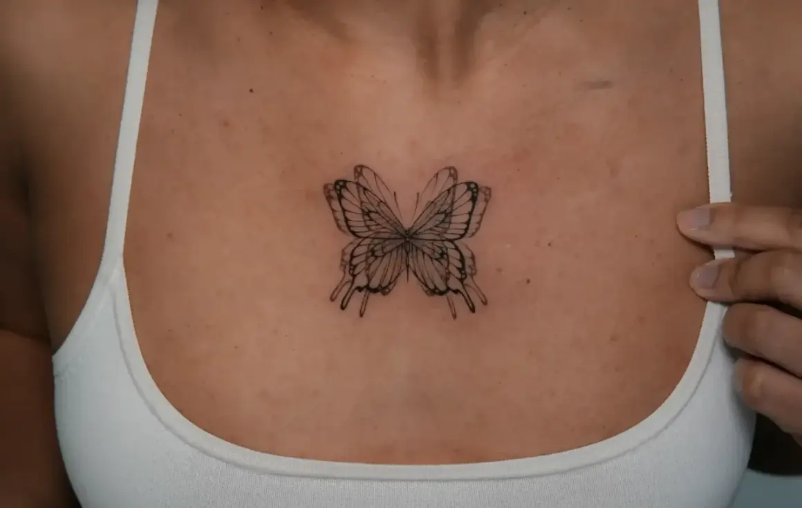 Zartes Fineline-Tattoo eines Schmetterlings zwischen den Brüsten, das wie ein filigranes Kunstwerk wirkt.