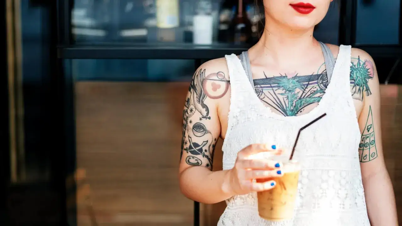 Frau mit vielen Tattoos, darunter ein auffälliges tattoo mit 16 auf dem Arm, hält einen Eiskaffee.