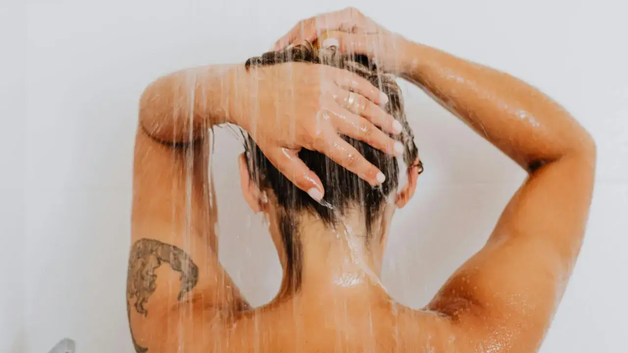 Eine Person mit einem frischen Tattoo wäscht sich unter der Dusche die Haare. Wasser rinnt über die Haut und das Kunstwerk.