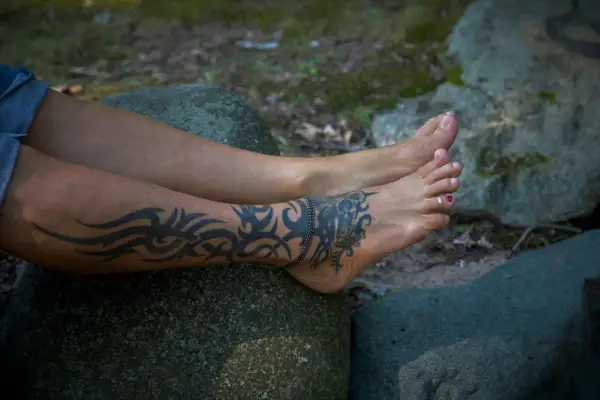 Beine mit einem Tribal-Tattoo, das sich über Wade und Fuß zieht. Das Tattoo kribbelt, als die Person auf einem Stein sitzt.