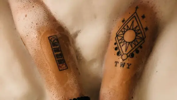 Zwei Arme mit Tattoos in Schaum gebadet. Ein Tattoo zeigt ein geometrisches Muster mit Sonne und Mond, das andere eine Reihe von Symbolen. PH neutrale Seife Tattoo-Pflege.