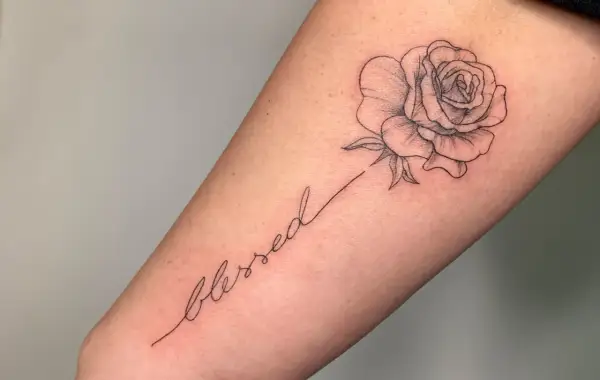 Ein zarter Fineline-Schriftzug "blessed" mit einer kleinen Rose als Tattoo.