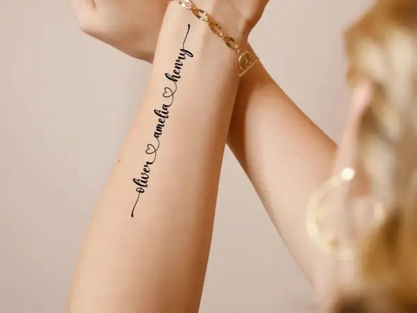 Ein zartes **Enkel Tattoo Namen** zeigt "oliver & amelia & henry" in geschwungener Schrift auf einem Arm, geschmückt mit einem goldenen Armband.