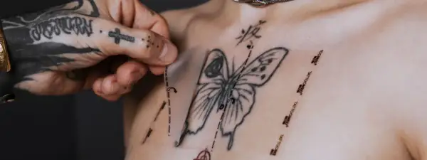 Ein Mann mit tätowierter Hand zieht eine Schutzfolie von einem frisch gestochenen Schmetterling-Tattoo auf der Brust ab. Pflege danach ist wichtig.