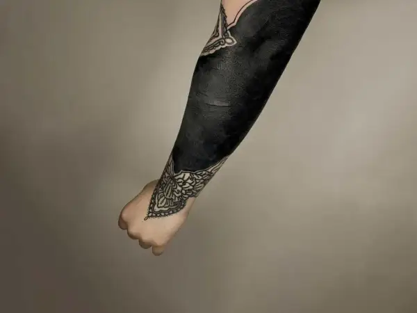 Ein Arm mit einem beeindruckenden **blackout tattoo**, das fast vollständig in tiefem Schwarz gehalten ist. Zarte Mandalas schmücken Handgelenk und Ellenbogen.