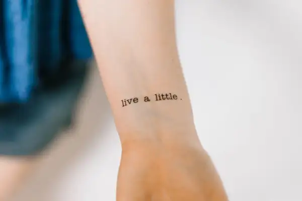 Ein Tattoo mit der Aufschrift "live a little" in Schreibmaschinenschrift auf einem Handgelenk.