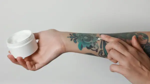 Pflege für dein Tattoo: Eine Hand hält eine Dose mit weißer Creme, während die andere Hand sie auf ein buntes Tattoo aufträgt. Kokosöl Tattoo-Pflege.