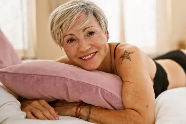 Lächelnde Frau mit kurzem blonden Haar und einem Stern-Tattoo auf der Schulter liegt auf einem Kissen.