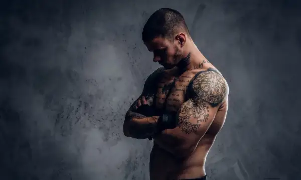 Ein muskulöser Mann mit vielen Tattoos, der seine Arme verschränkt. Sein Körperbau zeigt, dass er viel für Sport und Training tut.