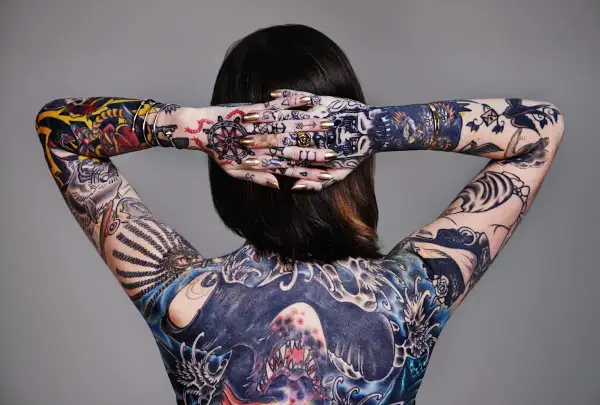 Rückenansicht einer Frau mit aufwendigen, abstrakten Tattoos, die ihren ganzen Körper bedecken. Goldene Nägel und Schmuck ergänzen die Kunstwerke.