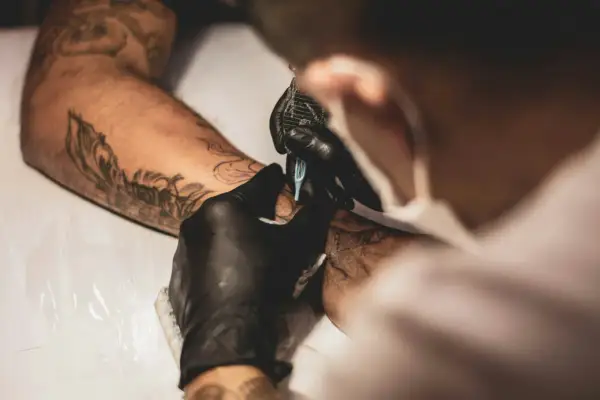 Tätowierer mit schwarzen Handschuhen arbeitet an einem Arm. Ein detailliertes Tattoo ohne Schattierung entsteht.